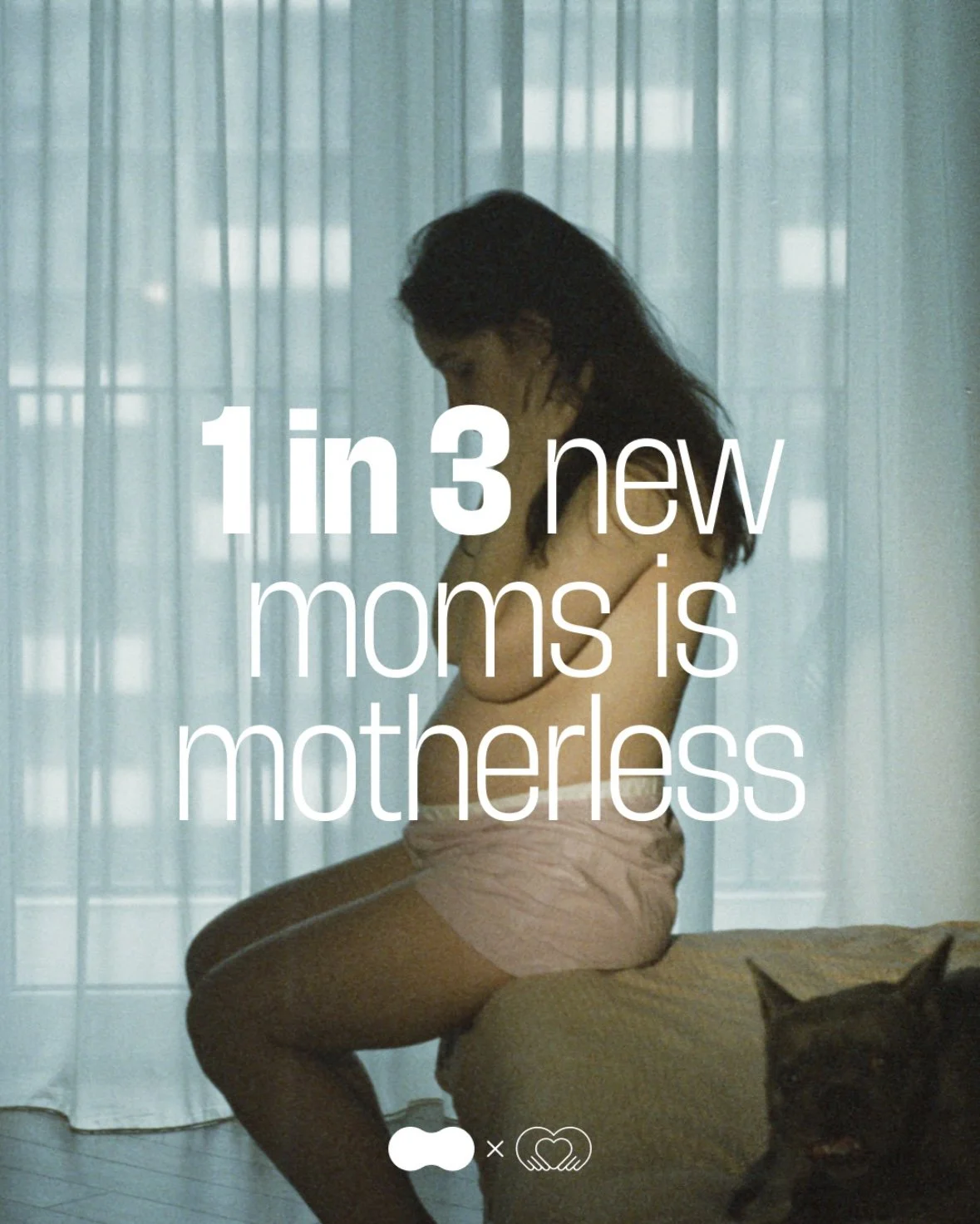 A woman sitting on a bed with her knees drawn to her chest, looking down, in front of a sheer curtain. There is a cat on the bed near her.