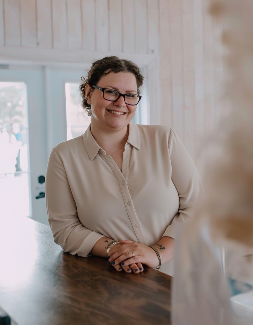 A woman with short curly hair, glasses, and tattoos on her arms is smiling and leaning on a wooden table inside a bright room with large windows.
