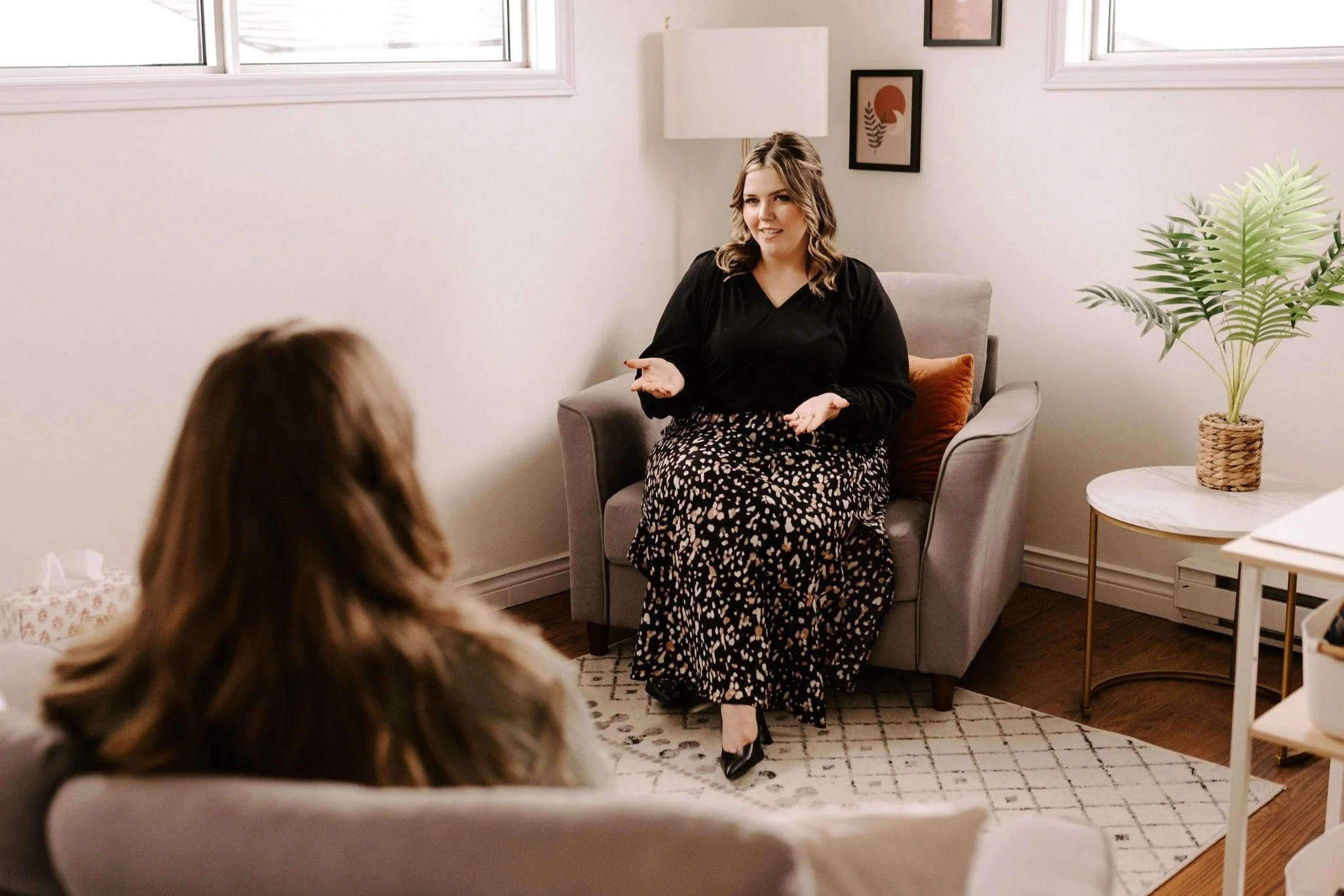 A woman in a black top and patterned skirt sits on an armchair, talking to another woman sitting with her back to the camera, in a cozy, well-lit room with framed art, a potted plant, and a side table.