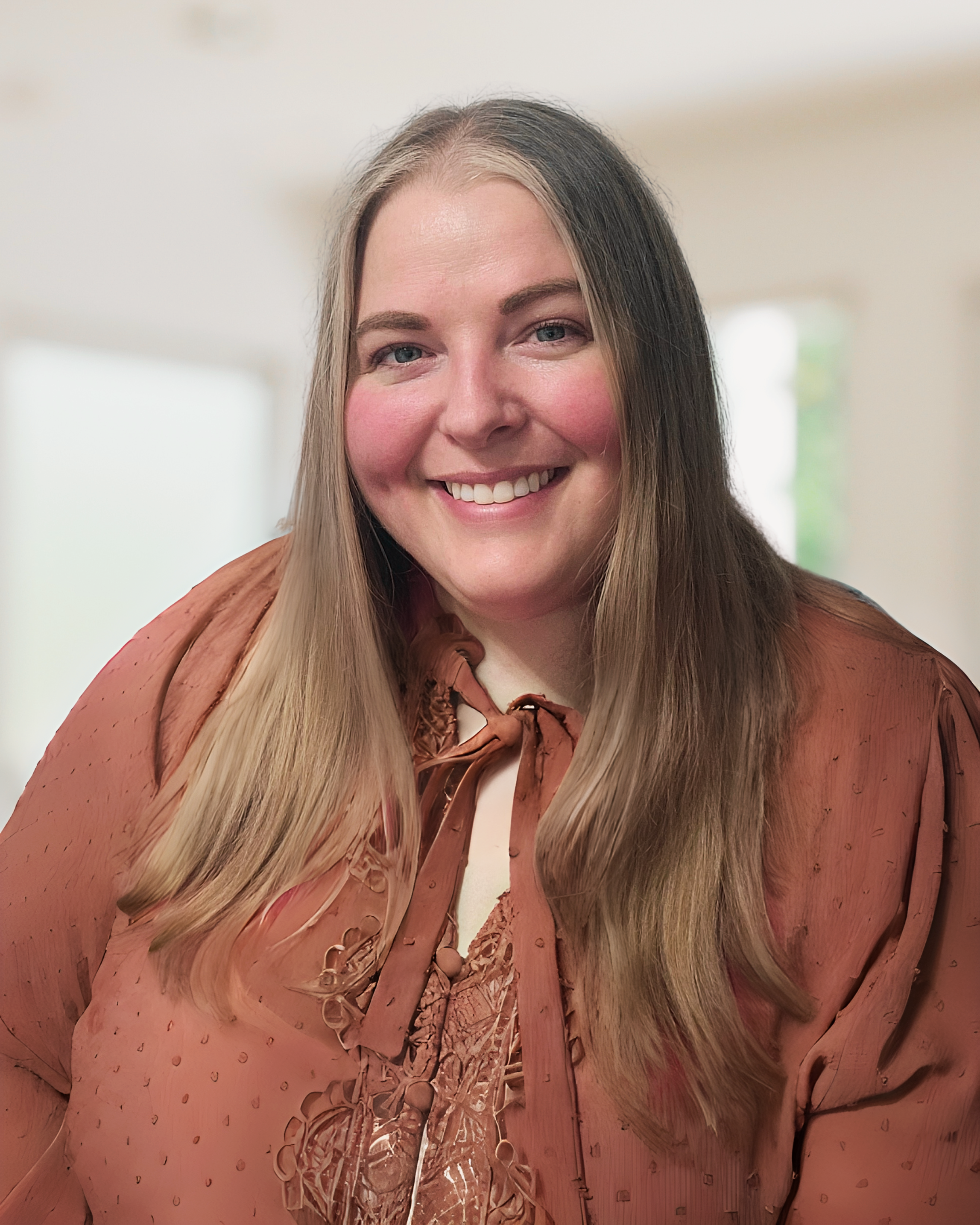 A woman with long, straight, light brown hair and a friendly smile wearing a light brown satin blouse with a bow tie.