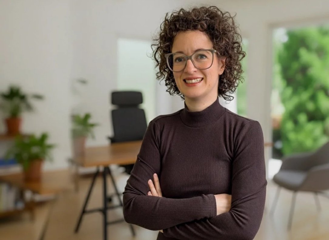 Smiling person with curly hair and glasses wearing a dark turtleneck and brown pants, standing in a modern, well-lit room.