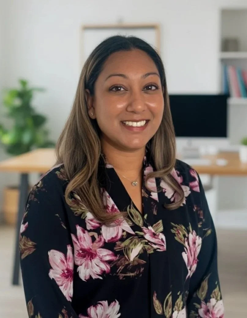 A smiling woman with shoulder-length hair, wearing a black floral blouse, standing indoors near windows with blurred greenery outside.