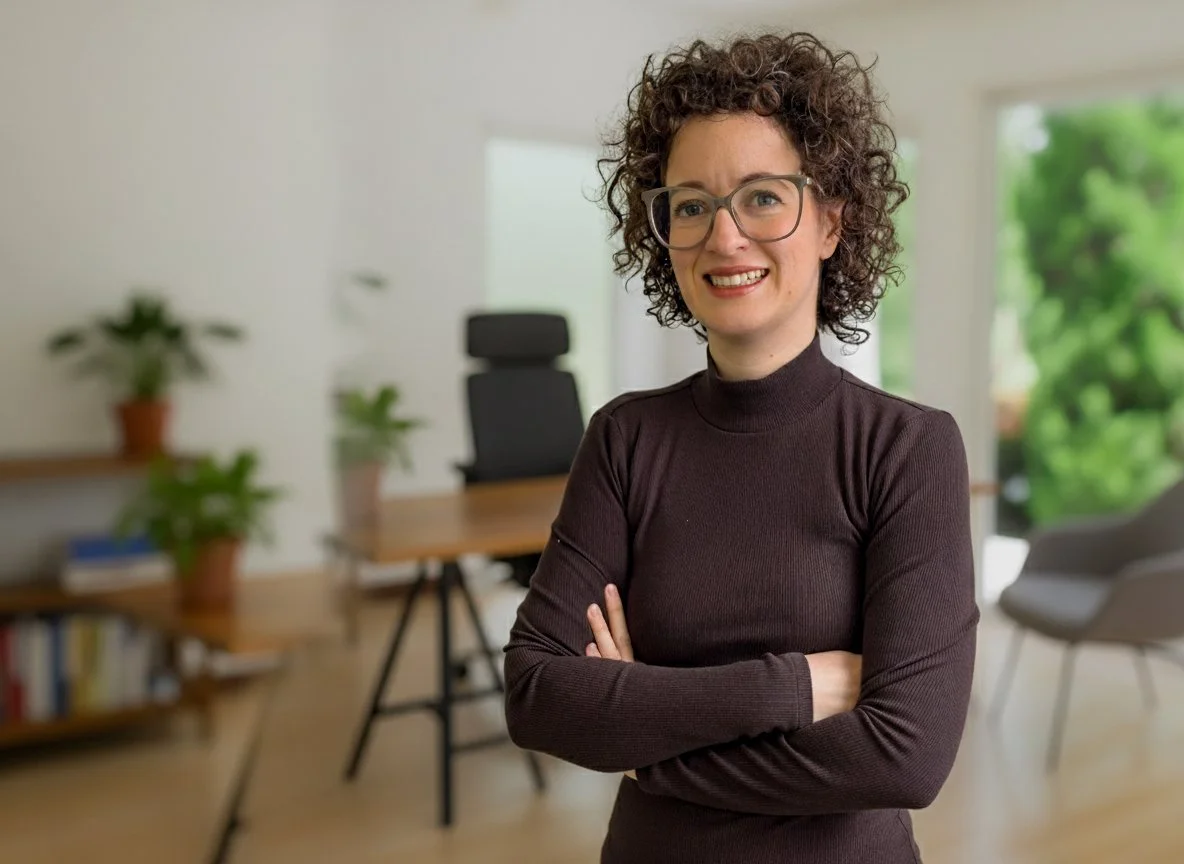 A woman with curly hair, glasses, and a dark turtleneck sweater crosses her arms and smiles at the camera.