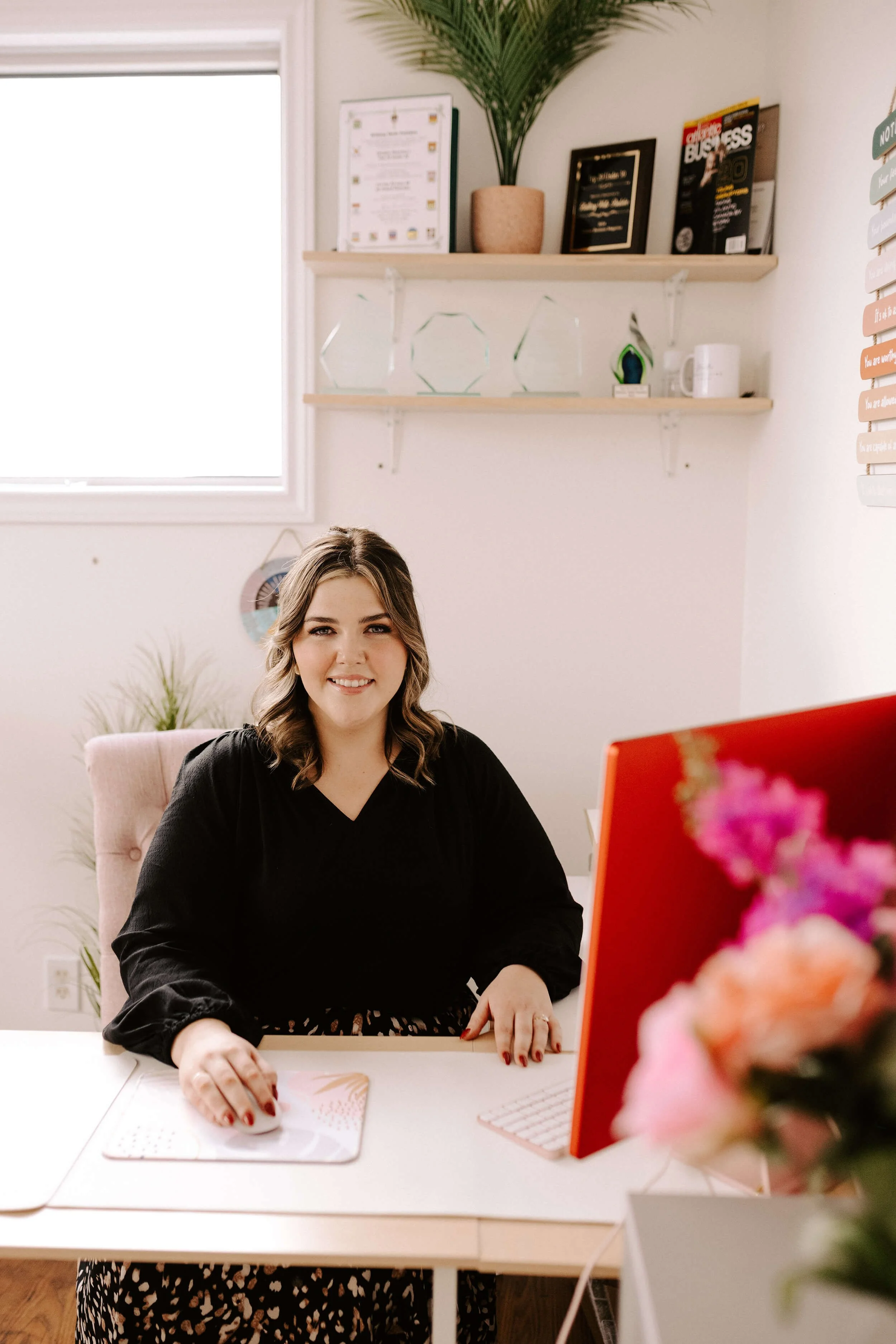 Woman sitting at a desk in an office, with a computer, mouse pad, and decorative shelves with awards and plant.