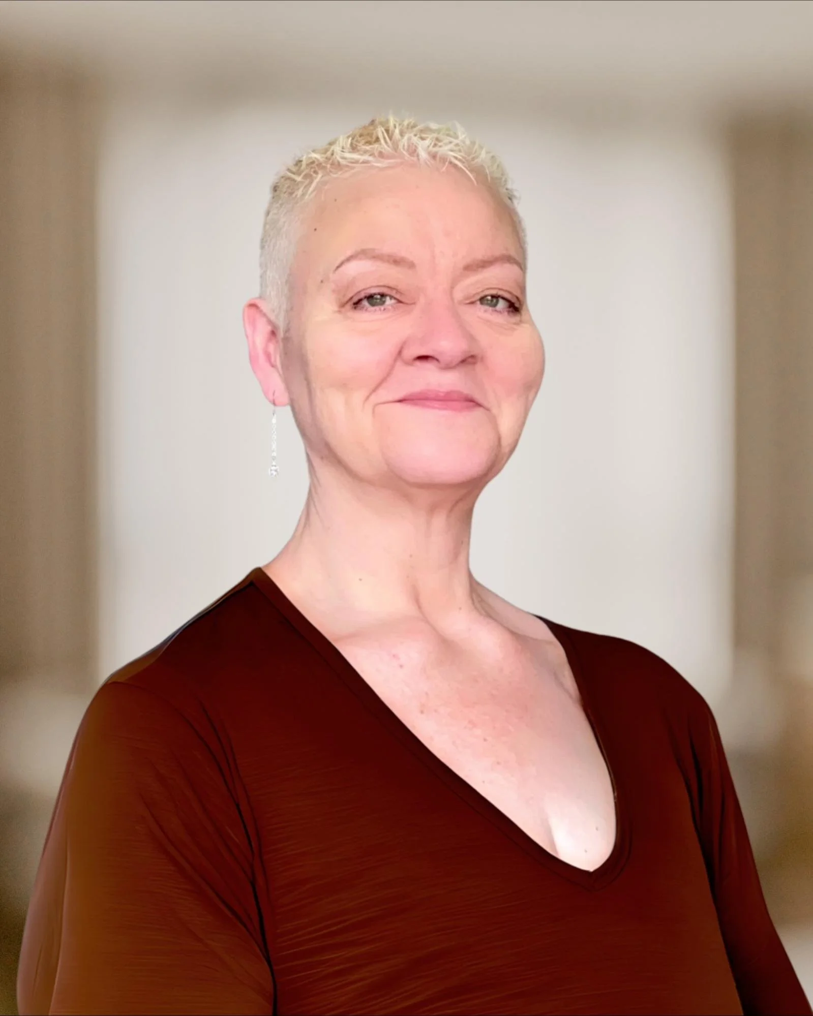 Person with short blonde hair wearing a brown V-neck top, smiling slightly, with a blurred background.