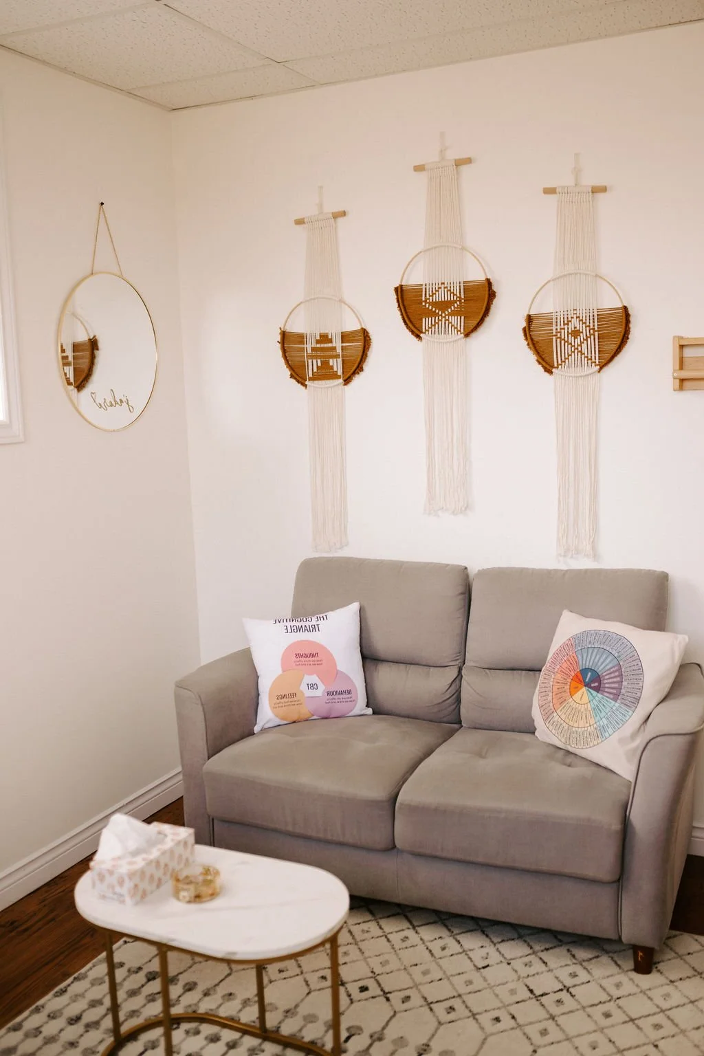 A cozy living room corner with a light gray sofa, decorative pillows, a white coffee table, wall hangings including a round mirror and three woven wall art pieces with macrame and wooden accents, and a patterned rug.