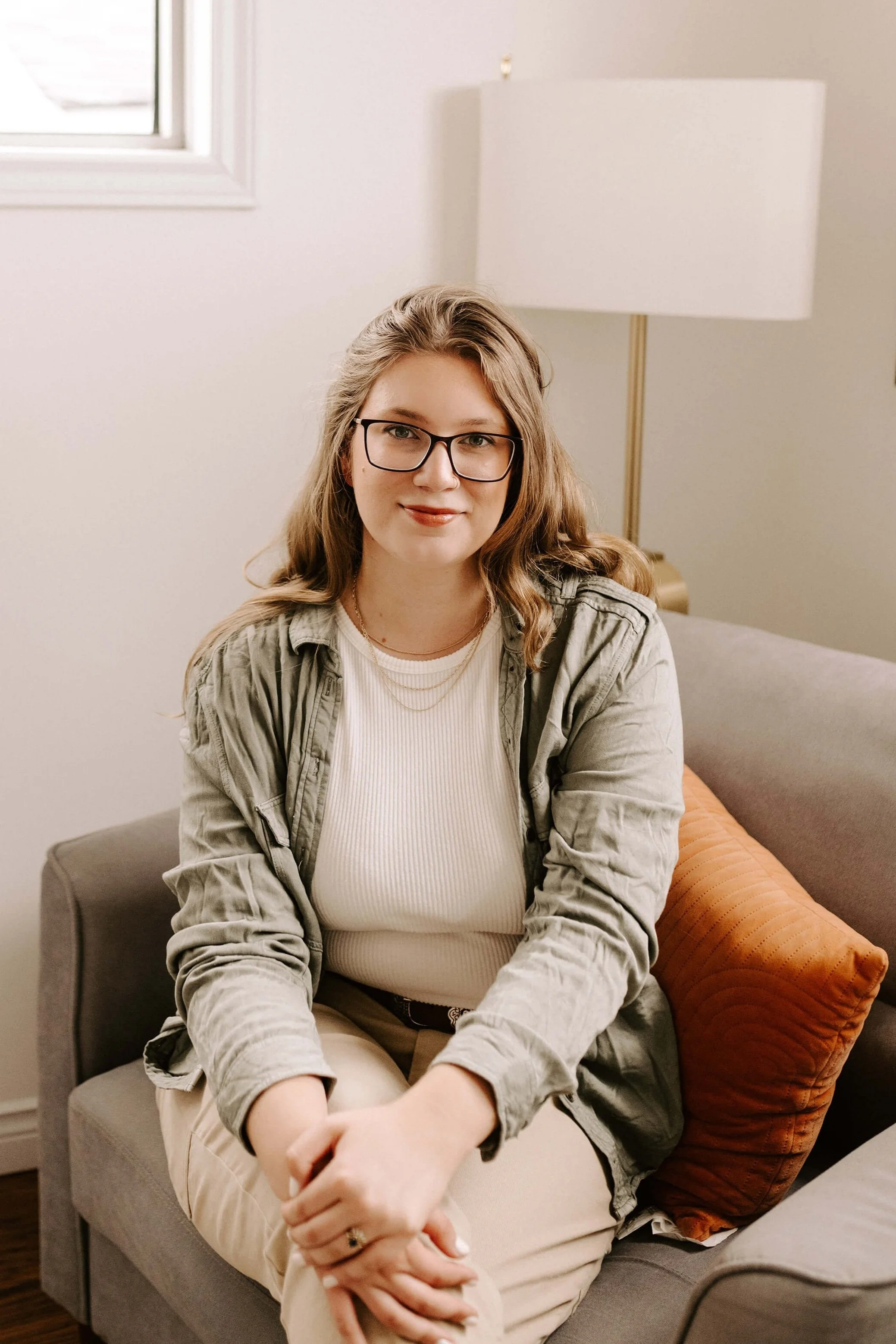A young woman with glasses and wavy blonde hair sitting on a gray couch, wearing a beige turtleneck, a green jacket, and beige pants, with an orange pillow behind her, in a cozy interior with a lamp and window.