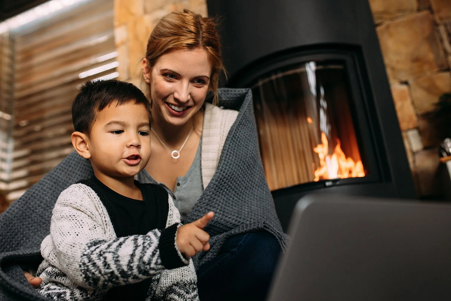 A woman and a young boy watching a computer in a cozy room with a fire burning in the fireplace.
