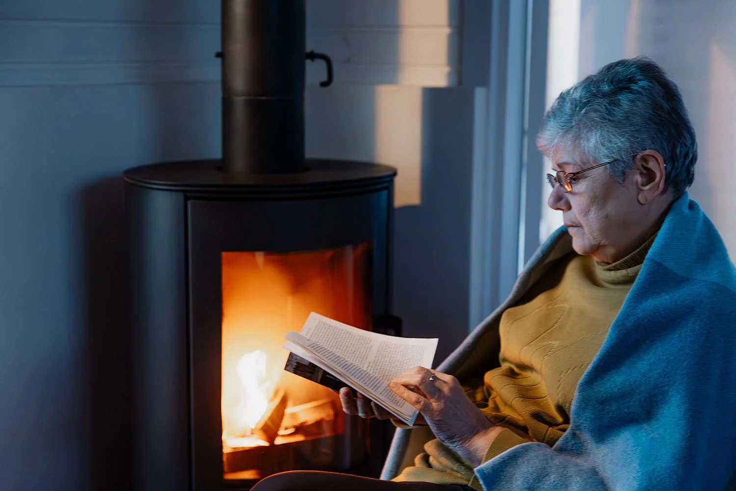An elderly woman sitting by a fireplace, reading a book, wrapped in a blanket.