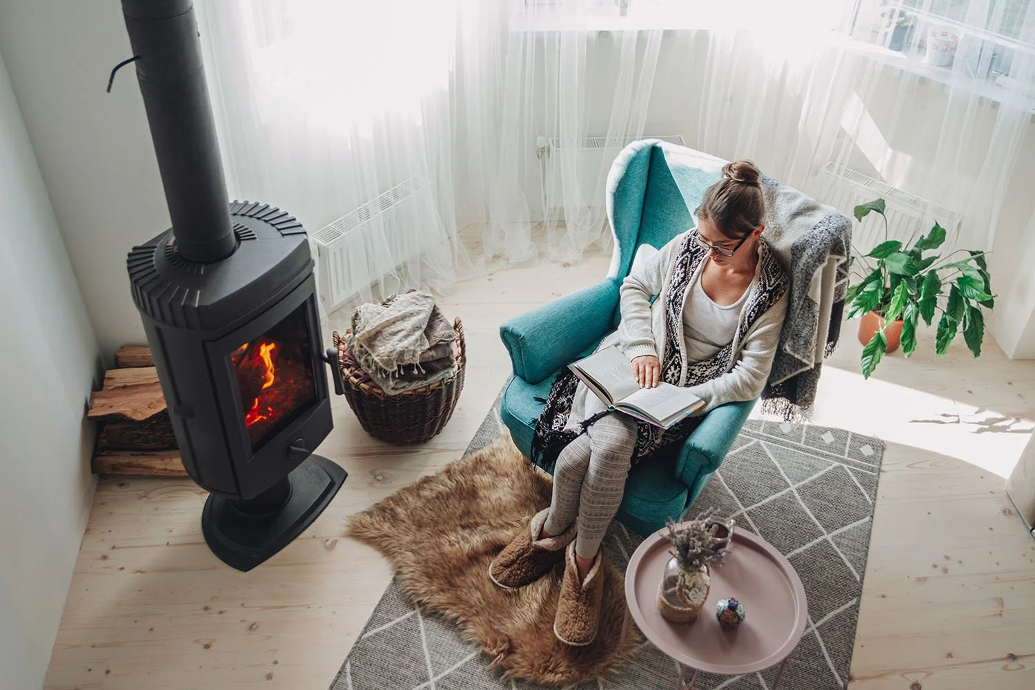 A woman with glasses reading a book while sitting in a teal armchair next to a wood stove in a cozy living room with natural light, a small round table with decorative items, a wicker basket with clothes, a plant, a fur rug, and a dark gray patterned