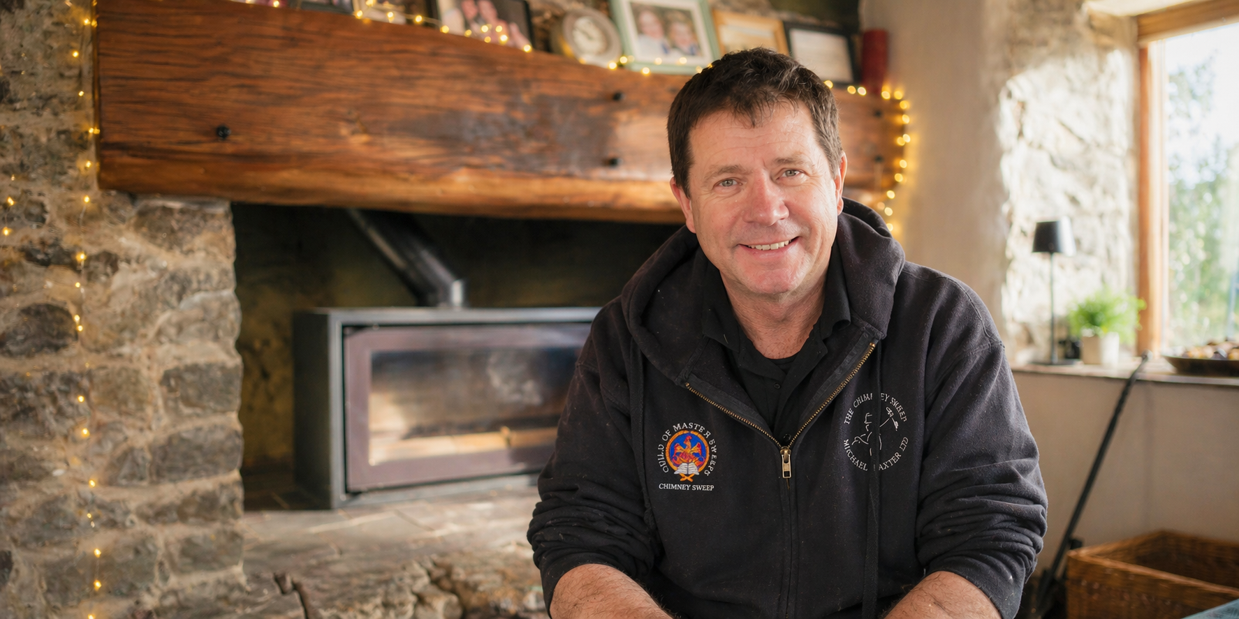 Michael Baxter The Chimney Sweep wearing a black hoodie with a logo on it, sitting in a cosy room with a stone fireplace, wooden mantel decorated with framed photos and string lights, and a window with natural light and plants inside.