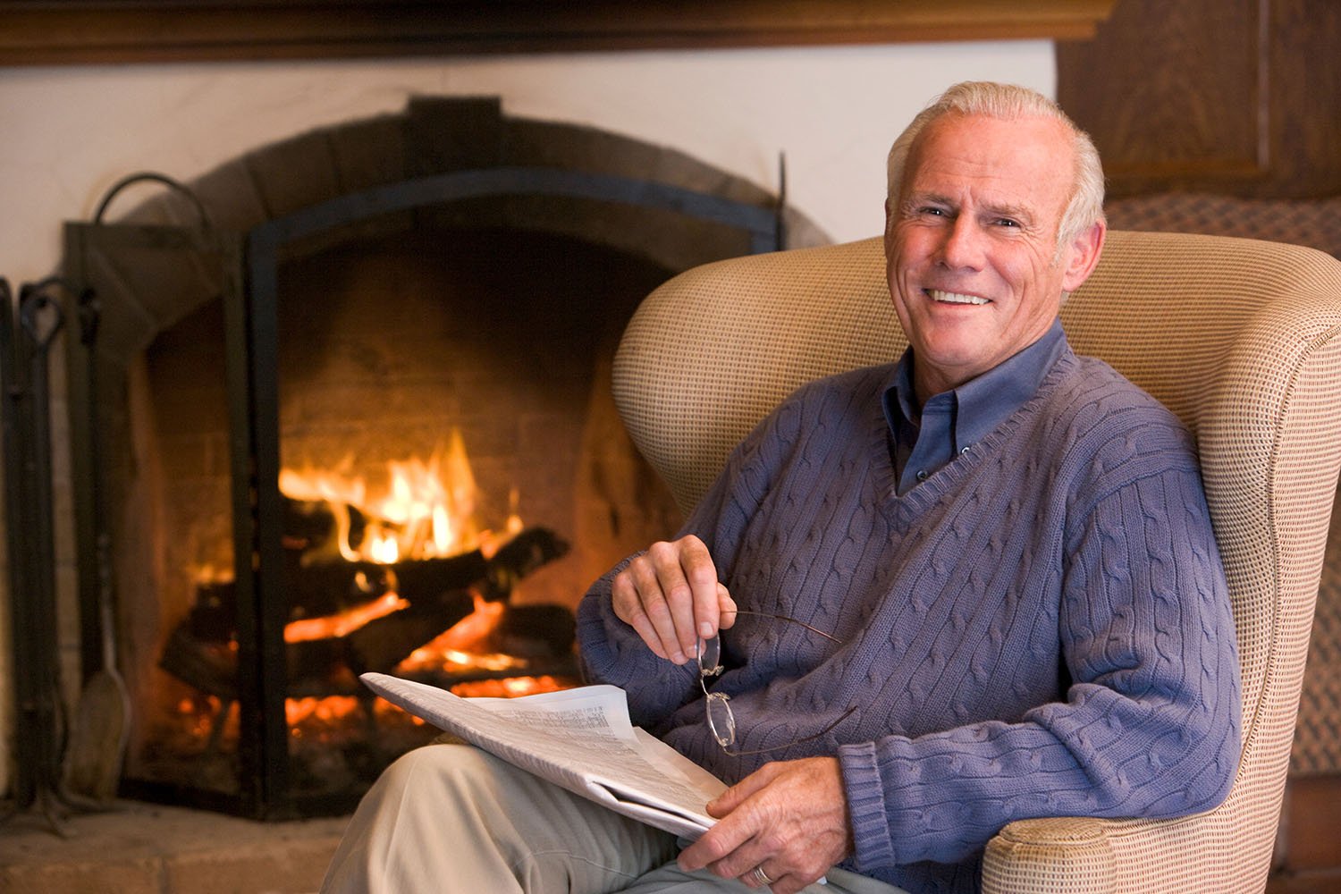 An elderly man sitting in a beige armchair reading a newspaper, smiling, with a lit fireplace in the background.