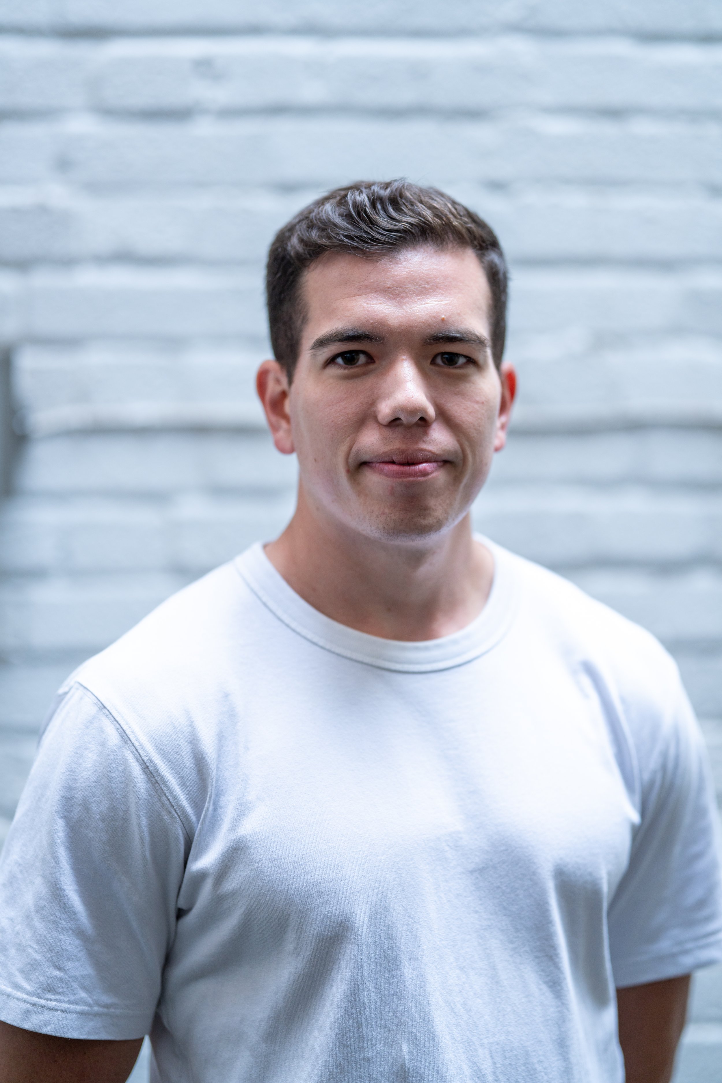 A young man with short dark hair wearing a plain white T-shirt, standing in front of a horizontal white brick wall.