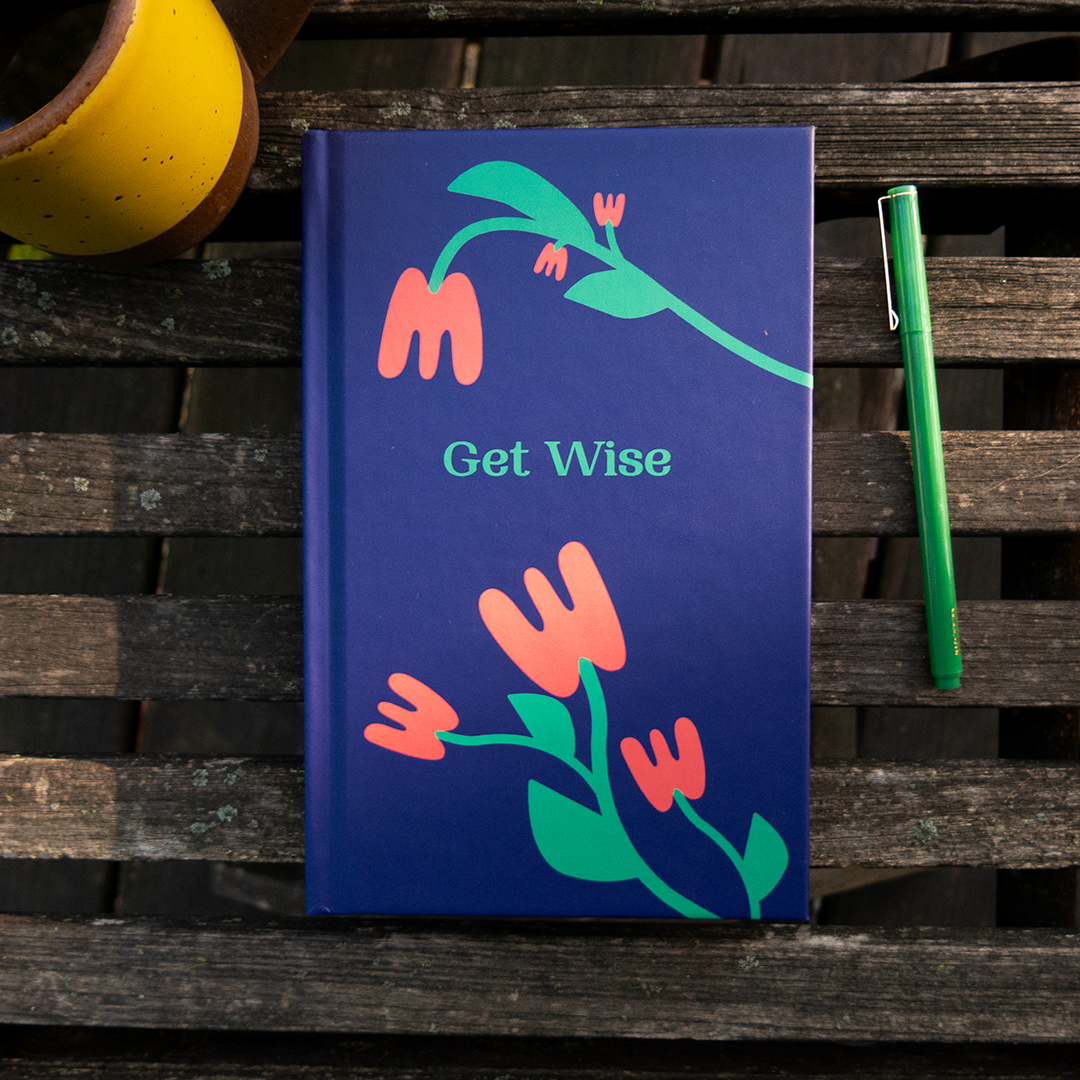 A navy blue hardcover book titled "Get Wise" with a colorful floral illustration on the cover, placed on a wooden surface next to a green pen and a yellow mug.
