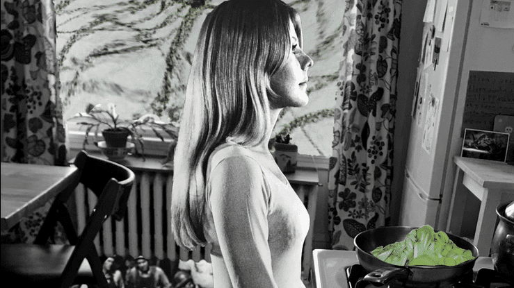 A woman with long hair standing in a kitchen, looking out a window with curtains, with plants and furniture in the background, and a pan with cooked greens on the stove.