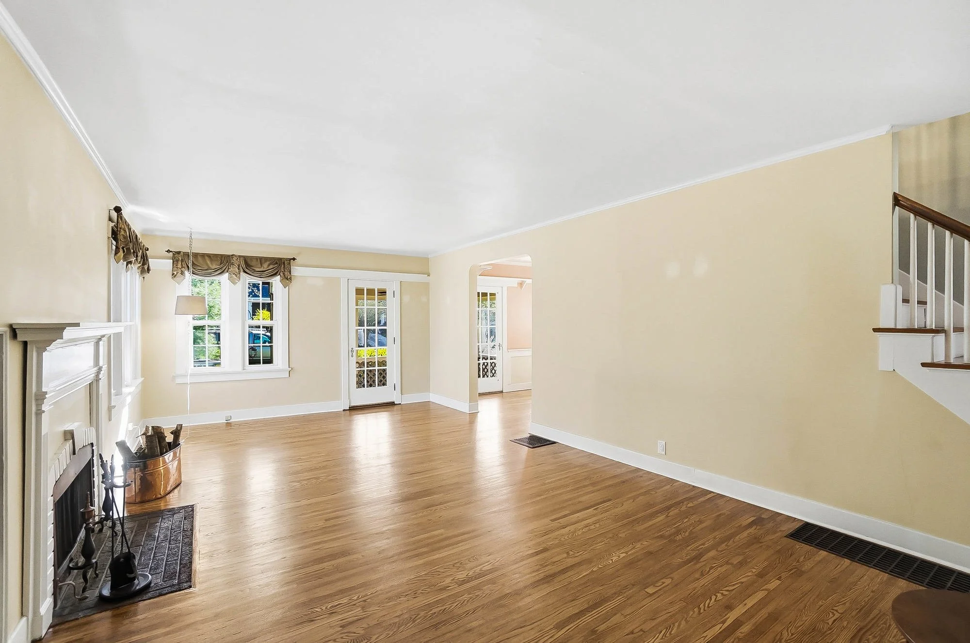 Empty living room with hardwood floors, cream-colored walls, two windows with curtains, and a door leading outside. There is a fireplace on the left and stairs on the right.
