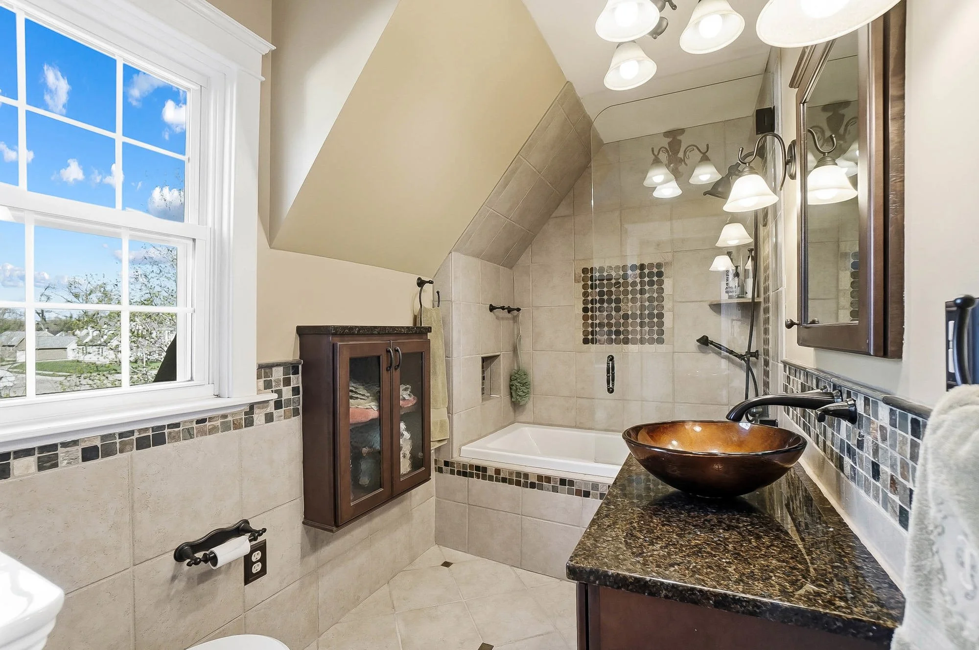 A bathroom with a large window showing a blue sky and clouds, a bathtub with beige tiles, a dark wooden cabinet, a granite countertop, a vessel sink, and multiple light fixtures with shades.