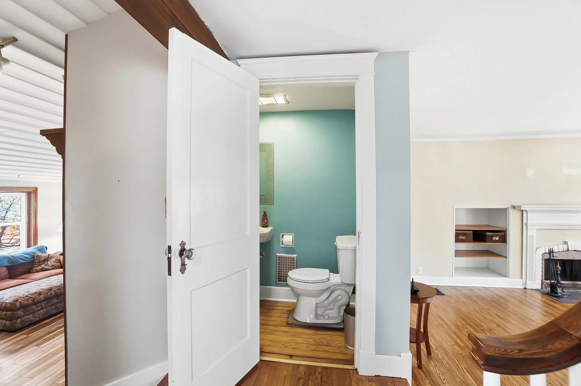 Bathroom with teal wall, toilet, small sink, and mirror, adjacent to living room with wooden flooring and beige wall.