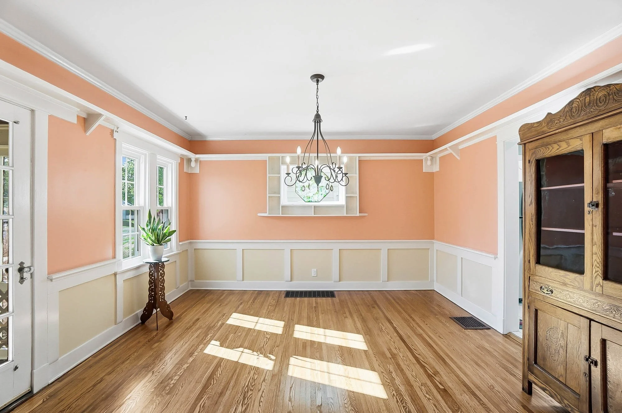 Empty dining room with peach and cream walls, hardwood floors, windows, a chandelier, an antique wooden cabinet, and a potted plant.