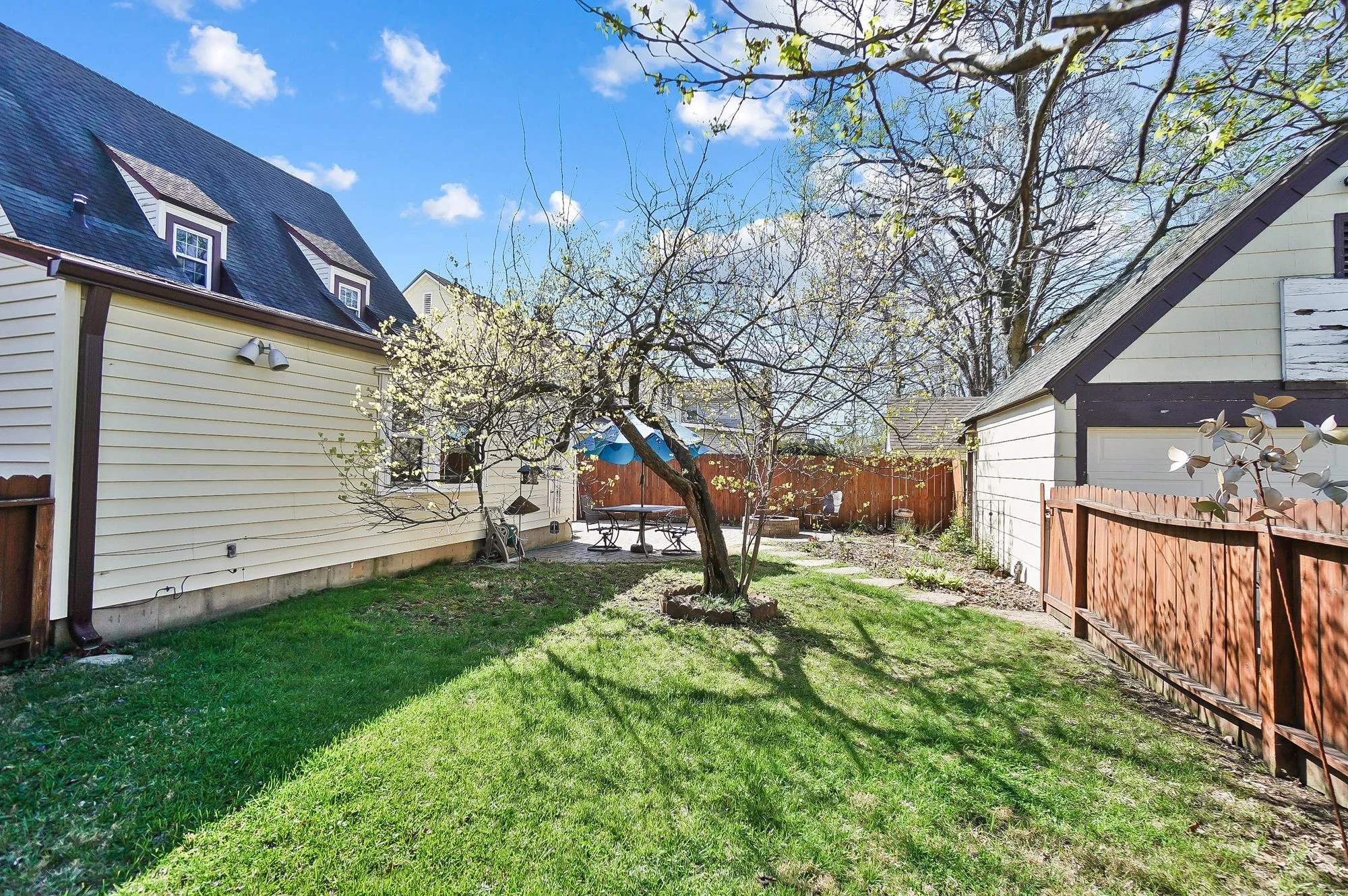 Backyard with a blooming tree, green grass, patio furniture, a blue umbrella, a fence, and neighboring houses under a partly cloudy sky.
