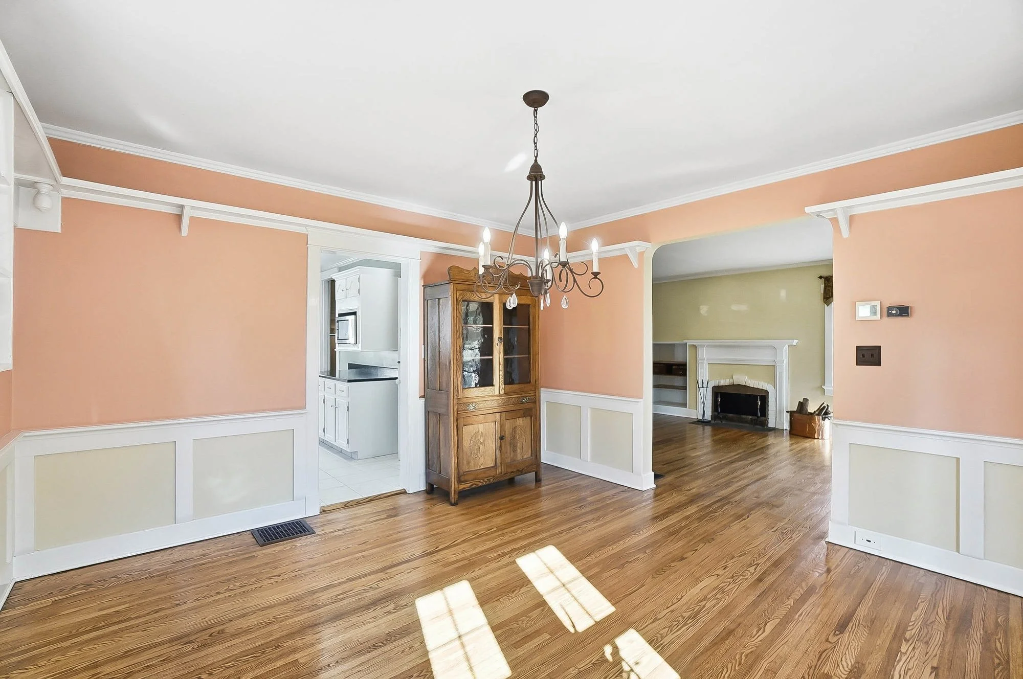 Empty dining room with hardwood floors, peach and cream walls, a chandelier, and a wooden china cabinet. The room connects to a kitchen and living room with a fireplace.