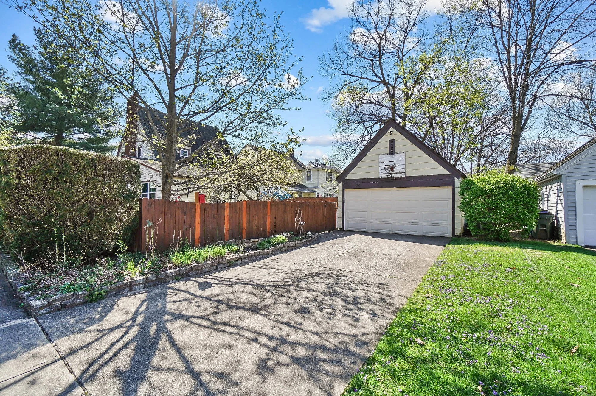 Residential backyard driveway with trees, a small shed with a basketball hoop on the front, a moss-covered hedge on the left, a fenced garden bed, and a grassy area to the right with small purple flowers.