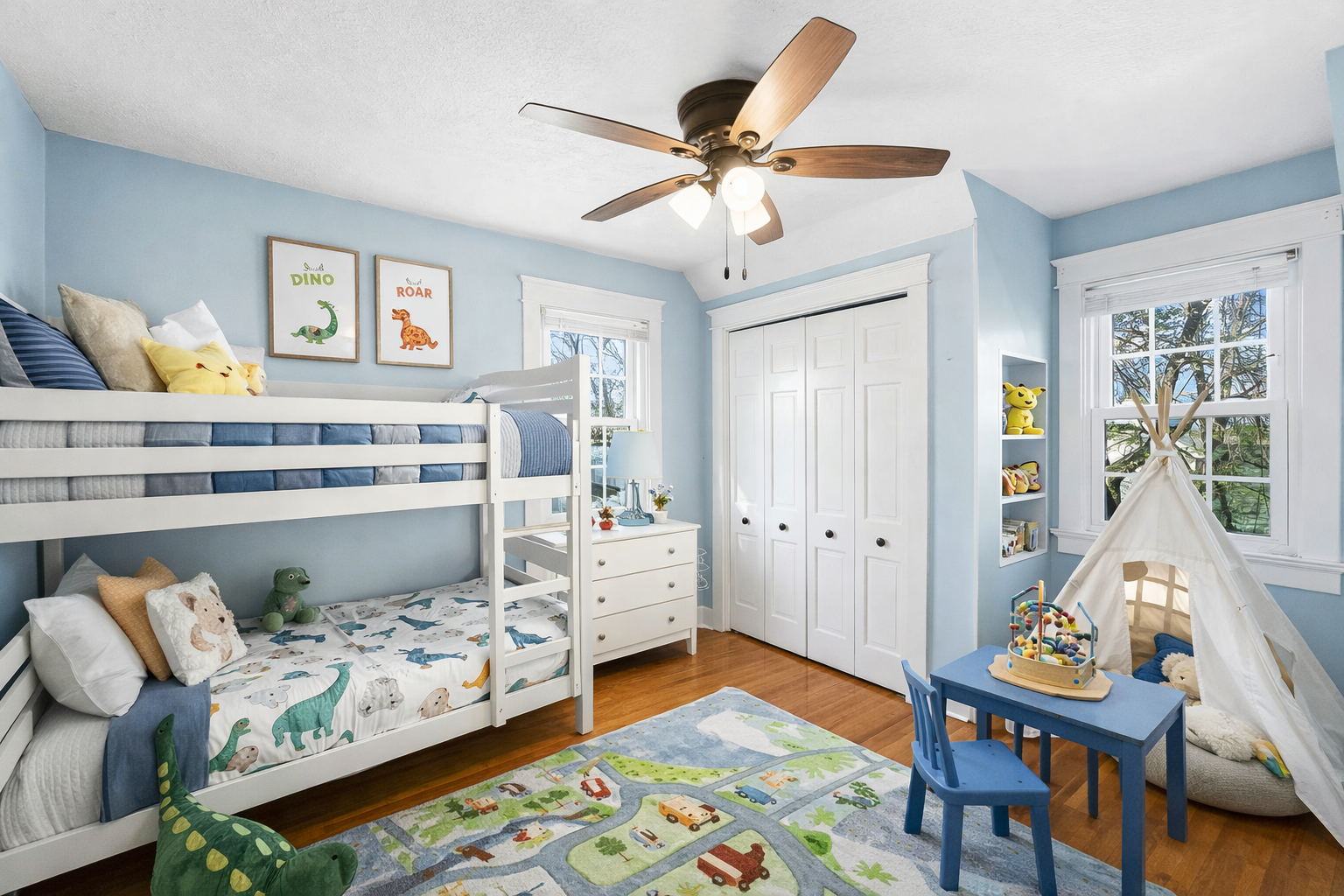 A children's bedroom with light blue walls, a white bunk bed with ocean-themed bedding, a small teepee tent, a blue table with a chair, a colorful play rug, and windows showing trees outside.