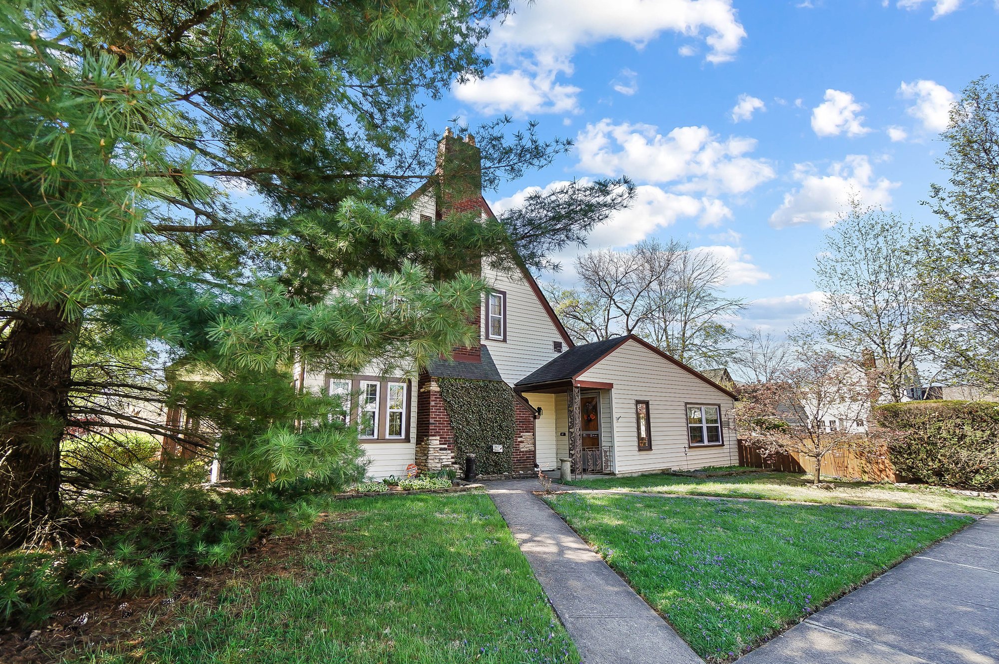 A house with a steep roof and brick chimney surrounded by trees and a front lawn on a sunny day.