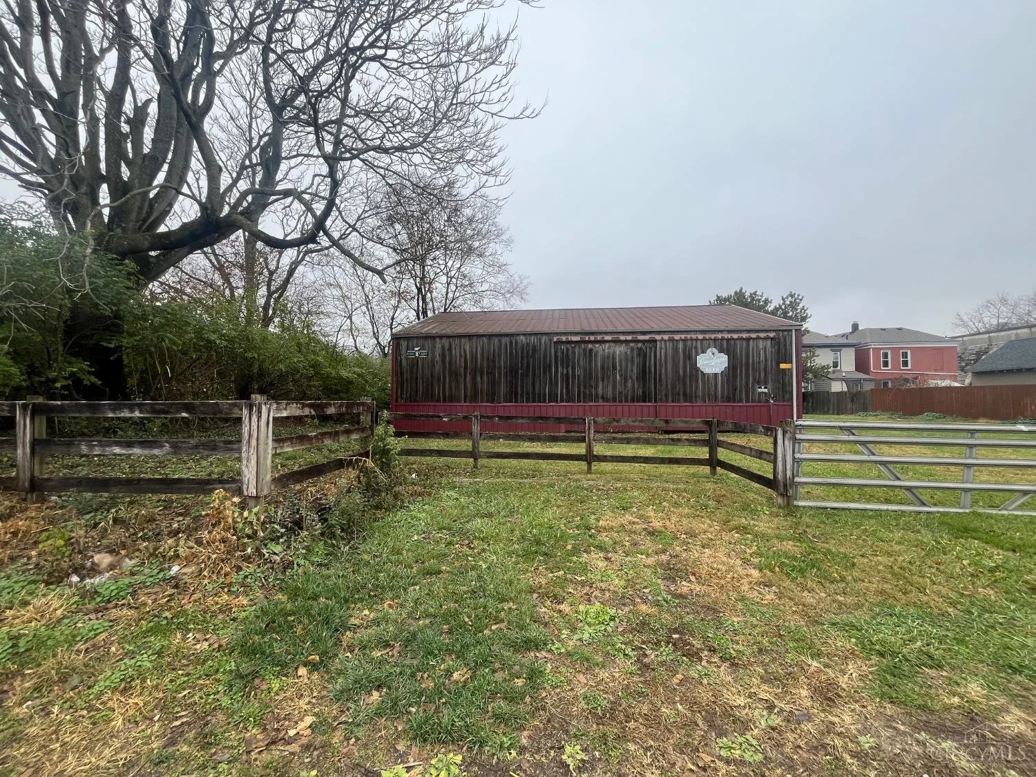 A backyard with a weathered wooden fence, a large leafless tree, a red barn, and neighboring houses under a cloudy sky.