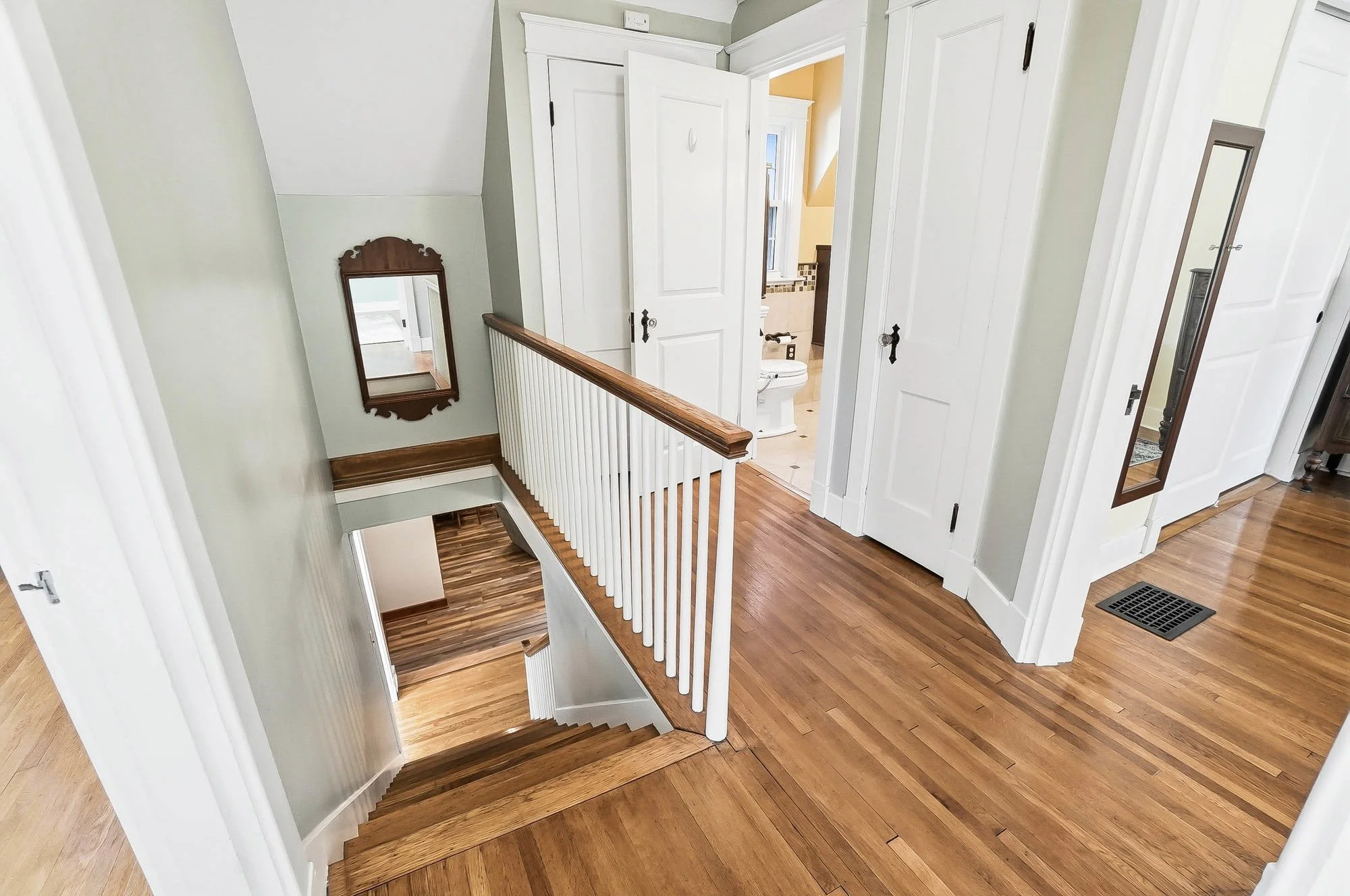 Interior view of a house top-floor landing with wooden flooring, white walls, a staircase with white spindles and wooden handrail leading downstairs, and open doors leading to a bathroom and closets.