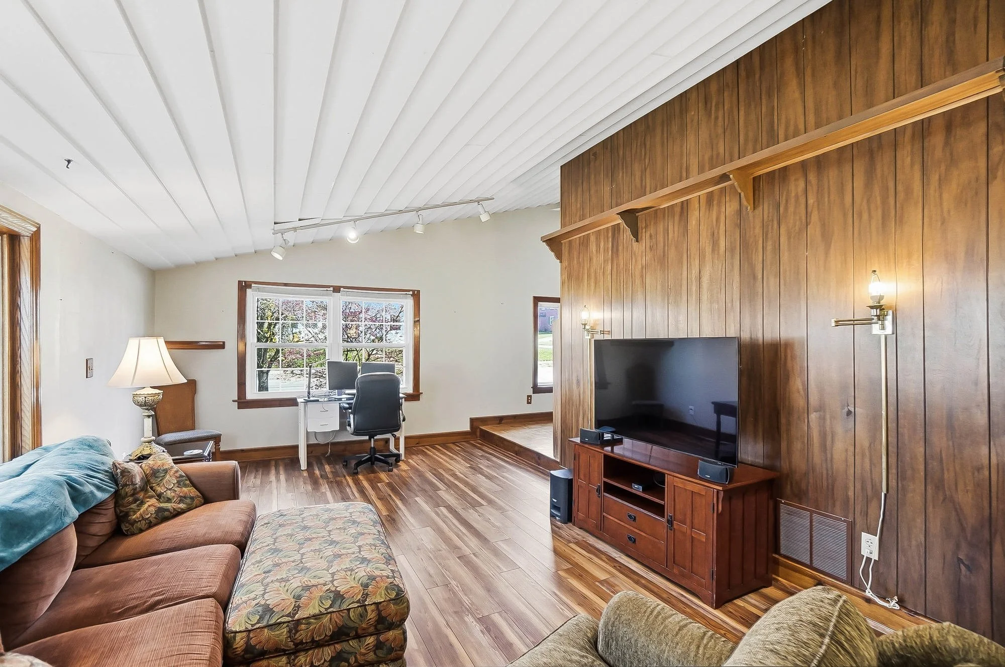 Living room with wooden floor, wood-paneled accent wall, a flat-screen TV on a wooden stand, a sofa with floral and plain cushions, a lamp, a window with a view of the yard, a desk with a chair, and wall-mounted lights.