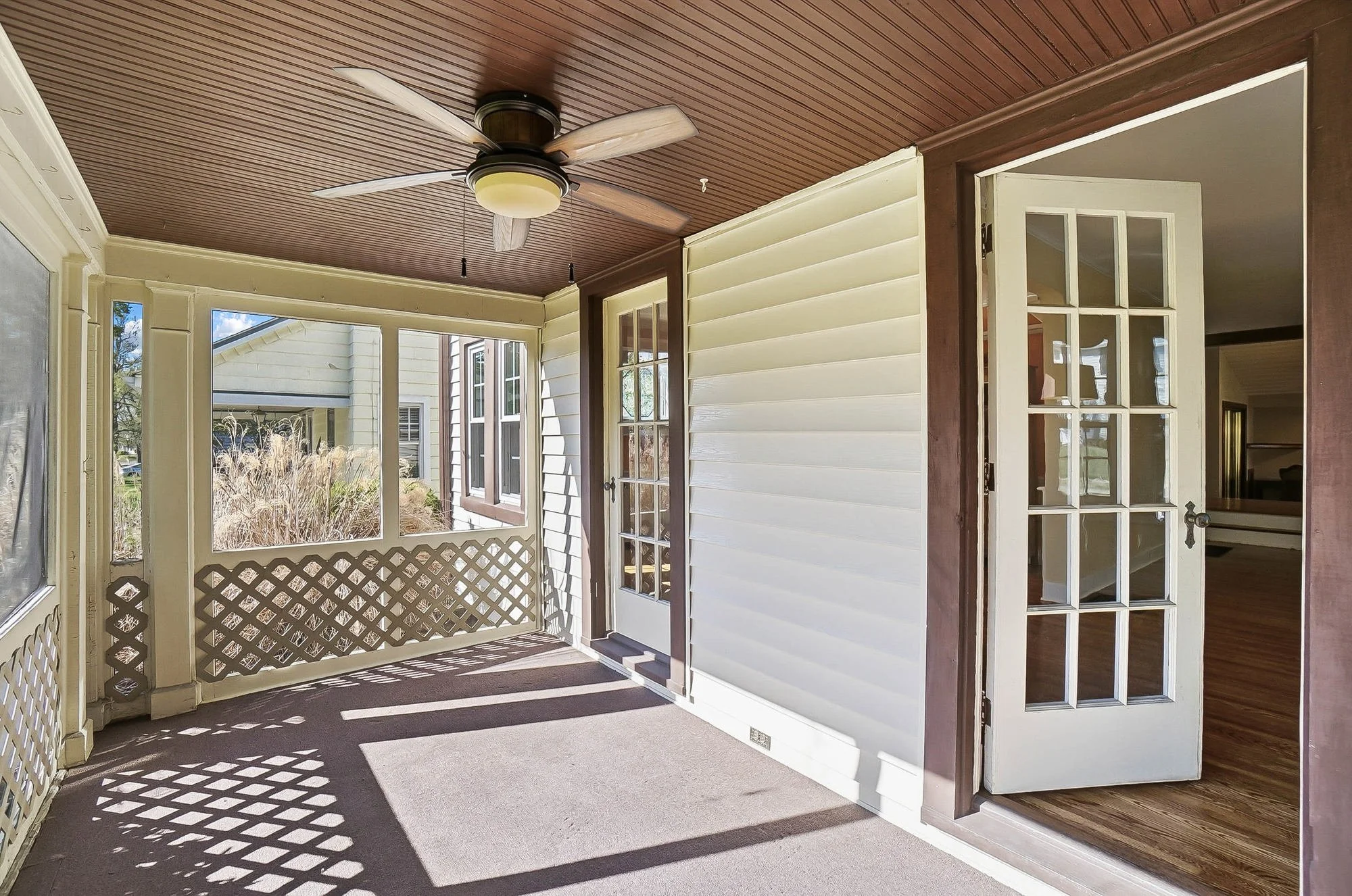 Sunlit enclosed porch with a ceiling fan, beige carpet, white siding, and a door leading to an indoor room. Outside view shows shrubs and part of the house.