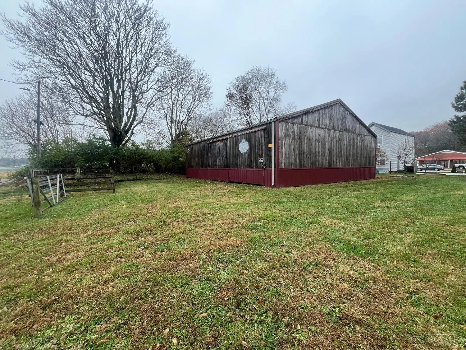 A grassy backyard with leafless trees, a wooden shed with a red base, and distant houses and cars under an overcast sky.