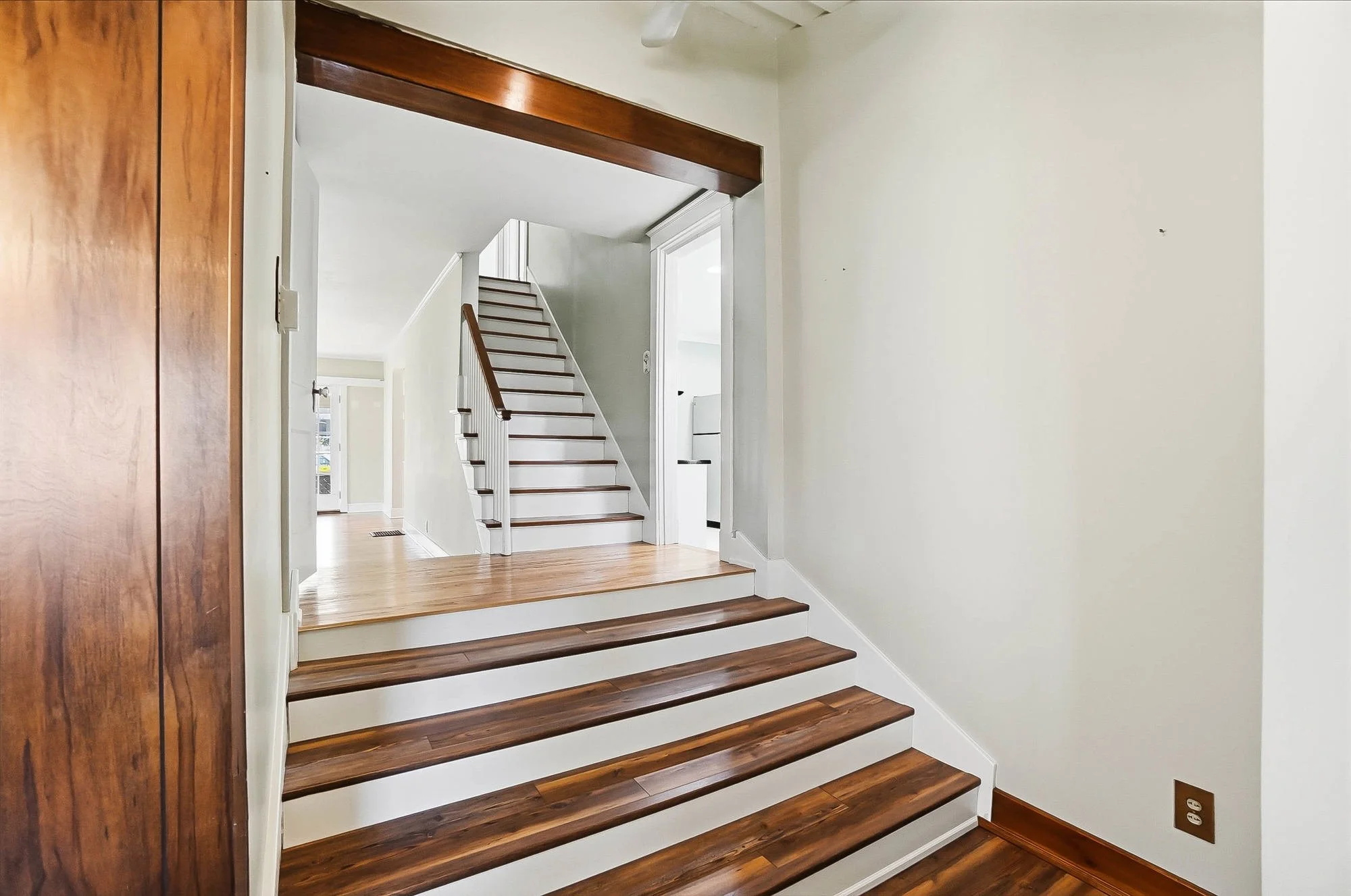 Interior view of a house with wooden stairs leading up to another floor. The staircase has white risers and wooden treads, with a white railing on the right side. The wall on the left side is painted a very light color, and there's a wall outlet visi