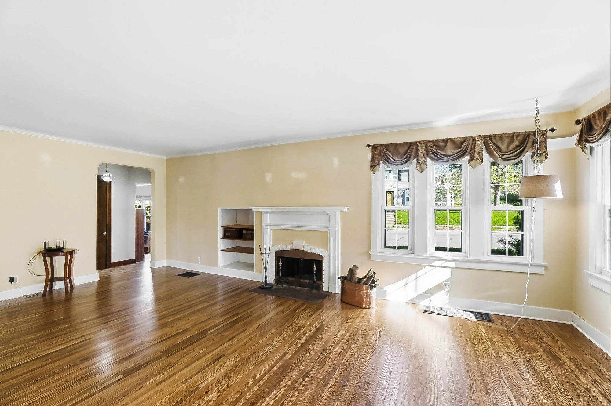 Empty living room with hardwood floors, cream walls, large window with draped curtains, fireplace, and simple furniture.