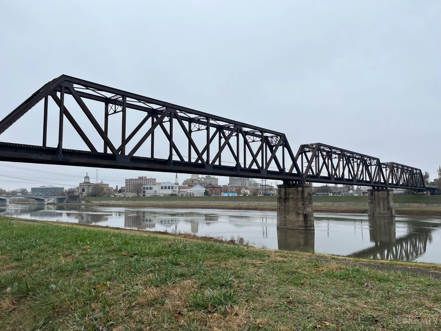 A steel train bridge over a river with city buildings in the background under an overcast sky.