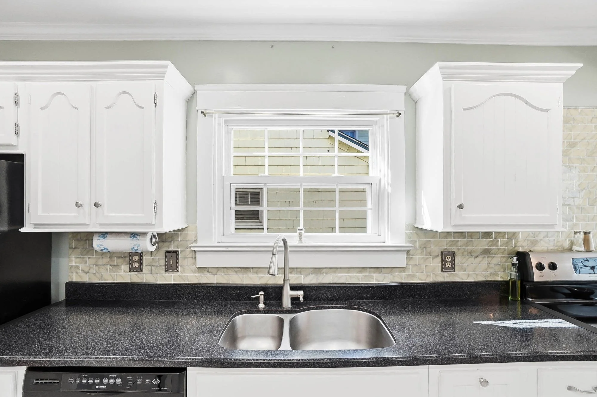 Kitchen sink with a window above, white cabinets on either side, black countertop, and appliances including a dishwasher and stove.