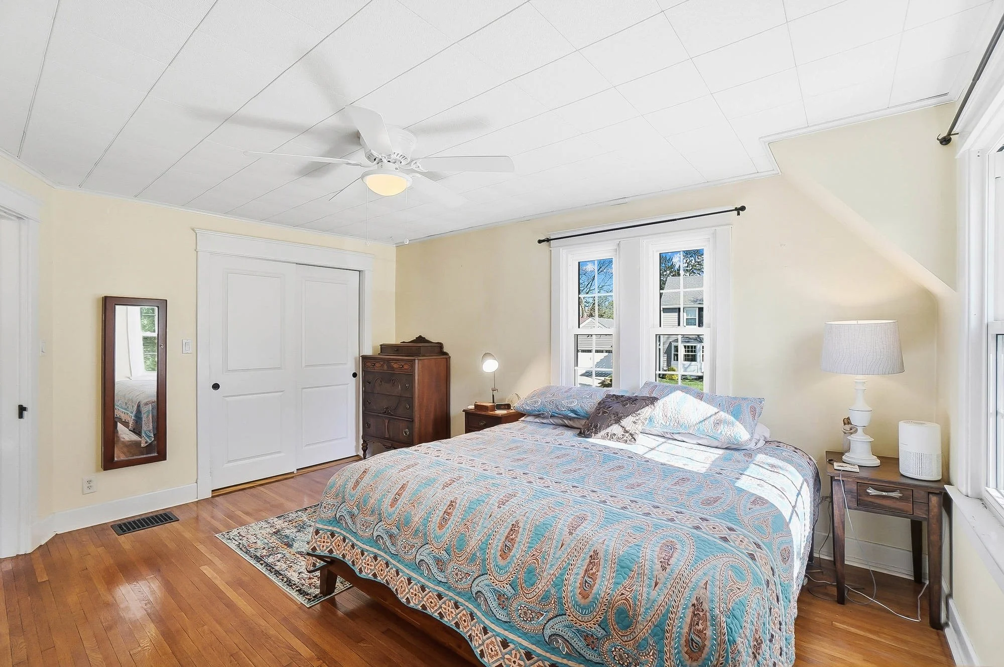 A bedroom with a bed, nightstands, a dresser, a full-length mirror, windows, a ceiling fan, and hardwood floors.