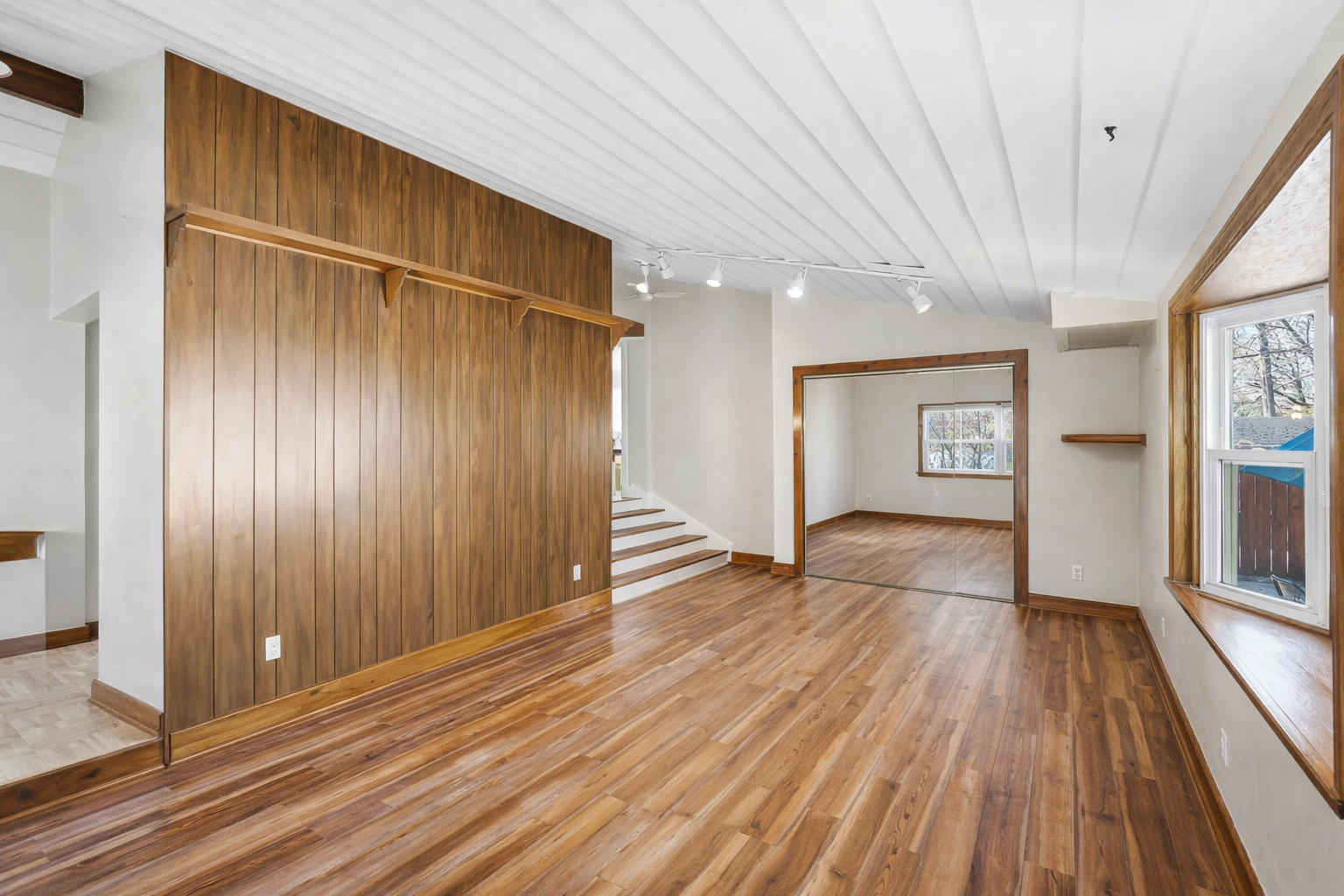 Empty living room with hardwood floors, wood paneling on one wall, a large window with a wood frame, and a mirrored wall.
