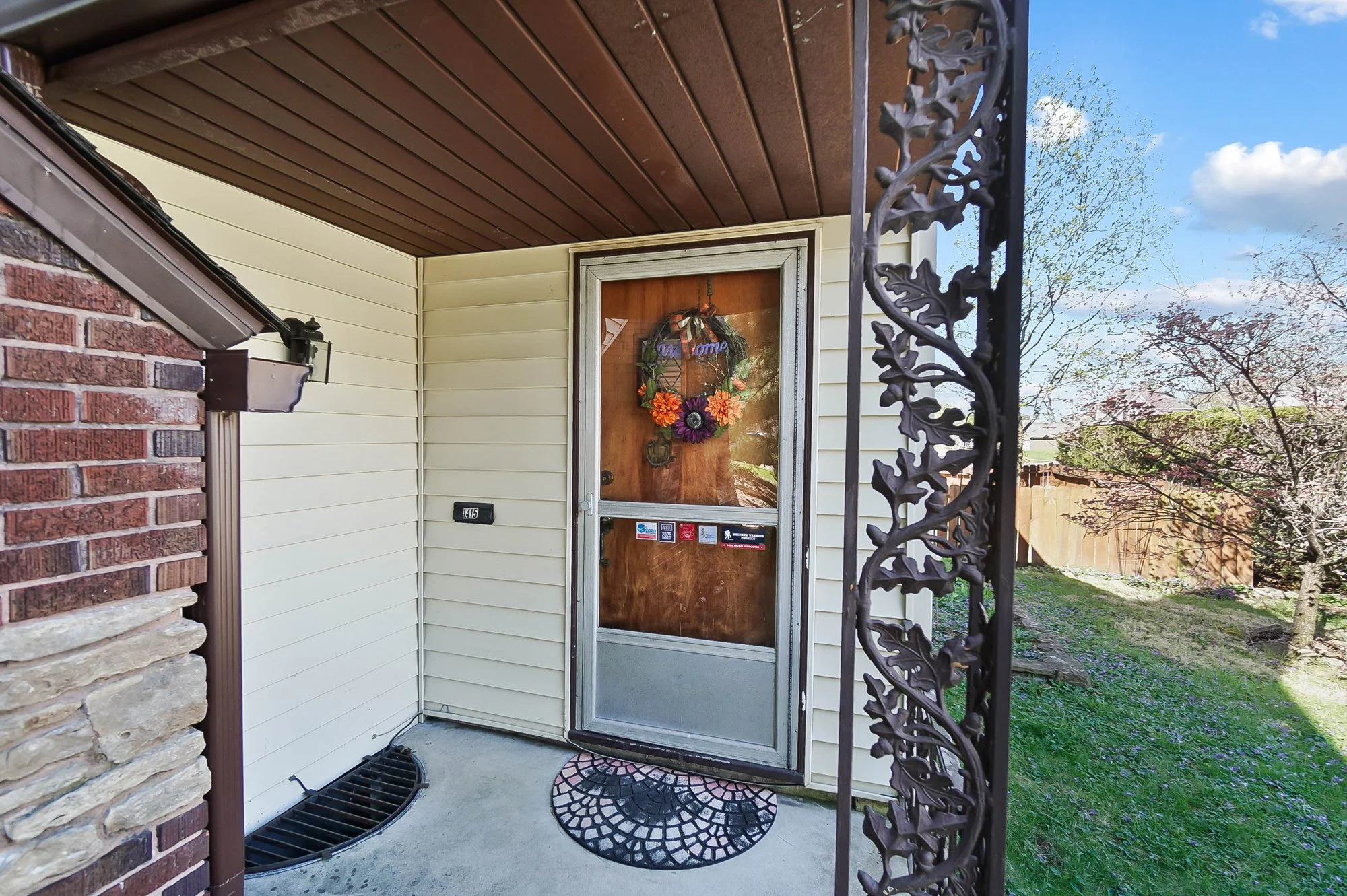 Front door of a house with a wreath hanging on a wooden door, a black metal decorative post on the right, and a small rainbow-colored welcome mat on the concrete porch.
