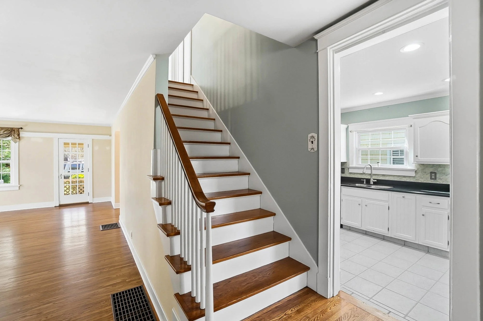 Interior view of a home showing wooden staircase, a kitchen with white cabinets, and a living room with hardwood floors and windows.