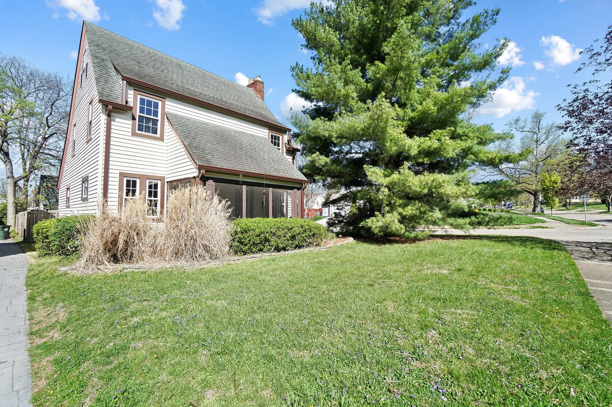 A house with white siding, multiple windows, and a grey roof, surrounded by a lawn, bushes, and a large evergreen tree under a blue sky with a few clouds.