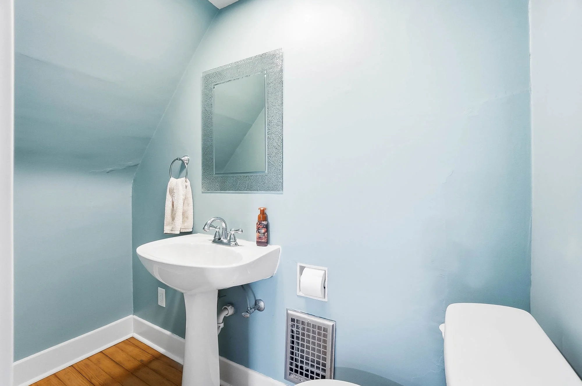 Small bathroom with light blue walls, white pedestal sink, mirror, towel ring with a towel, bottle of hand soap, toilet paper dispenser, vent on the wall, and part of a toilet visible.