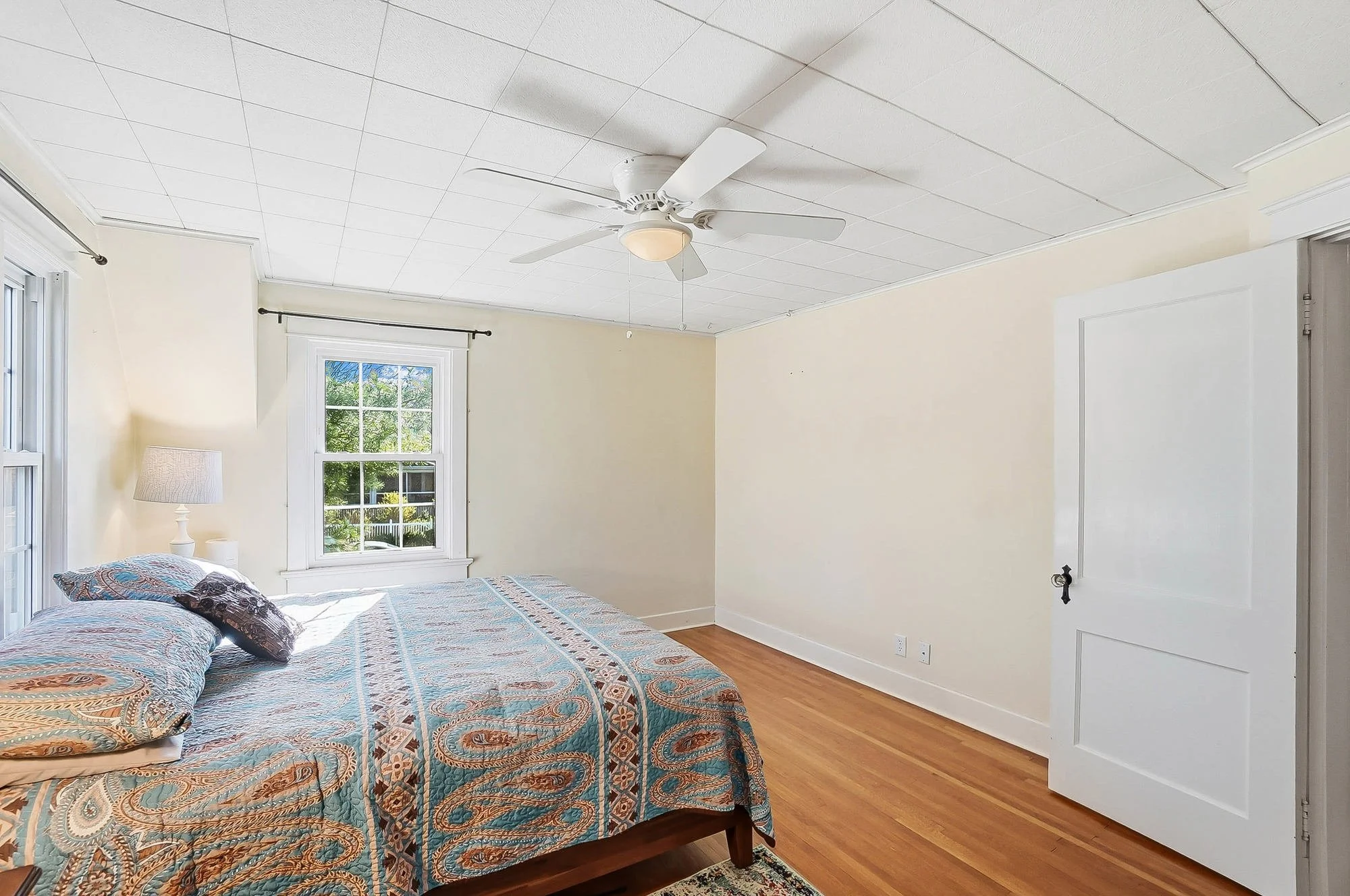 A bedroom with a bed covered in a patterned quilt, two pillows, a window with a view of greenery, a ceiling fan with a light, and a door on the right side.