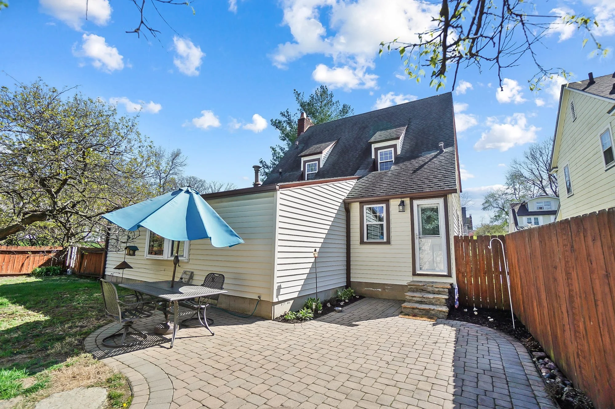Backyard patio with a round table, four chairs, and a blue umbrella, surrounded by a wooden fence and trees, with a house in the background under a blue sky with clouds.