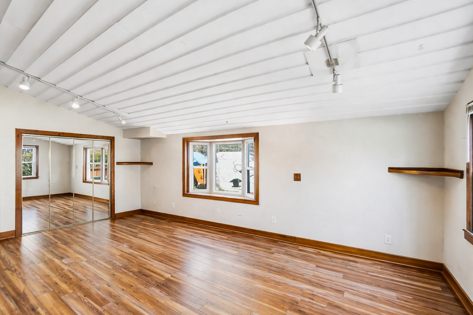 Empty room with hardwood floors, white walls, wooden window frames, a mirrored closet with sliding doors, and two small wall-mounted shelves.