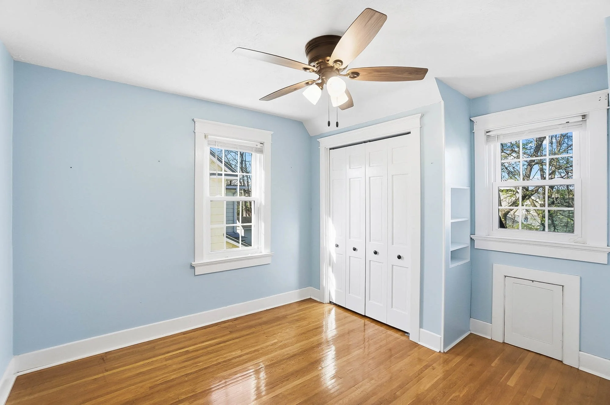 Empty room with light blue walls, hardwood floor, two windows, a ceiling fan, and a closet with white doors.