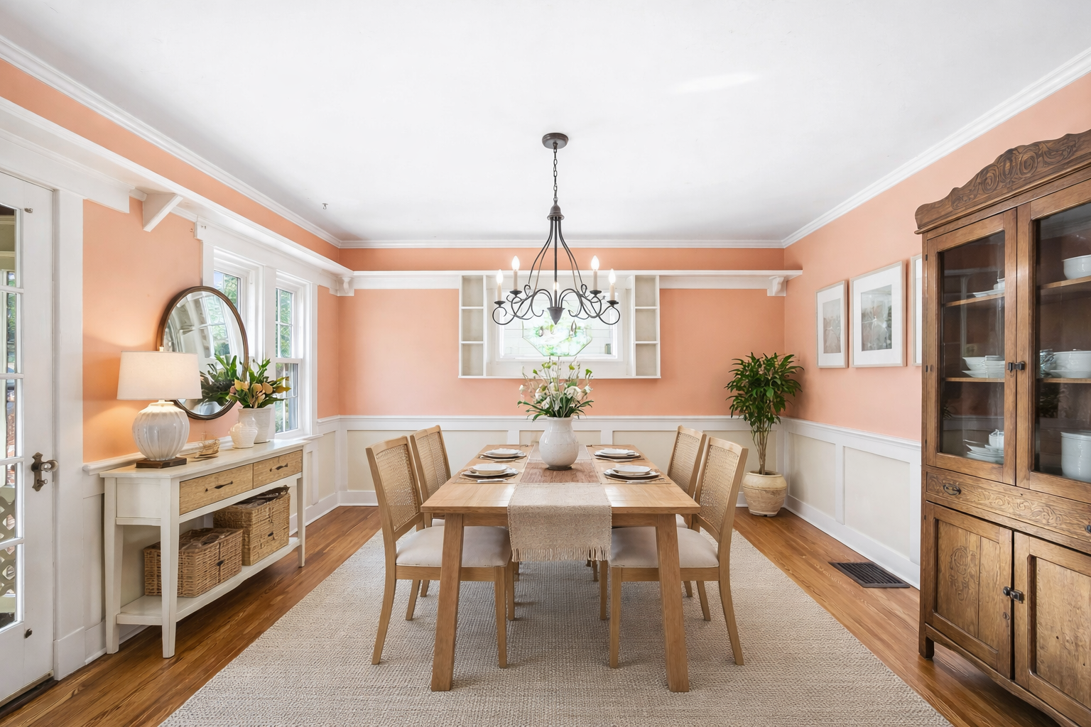A dining room with peach walls, a wooden dining table set with six plates, a white vase with flowers, and a chandelier hanging above. There are three windows with white trim, a sideboard with a lamp and plants, and a wooden china cabinet with dishes 