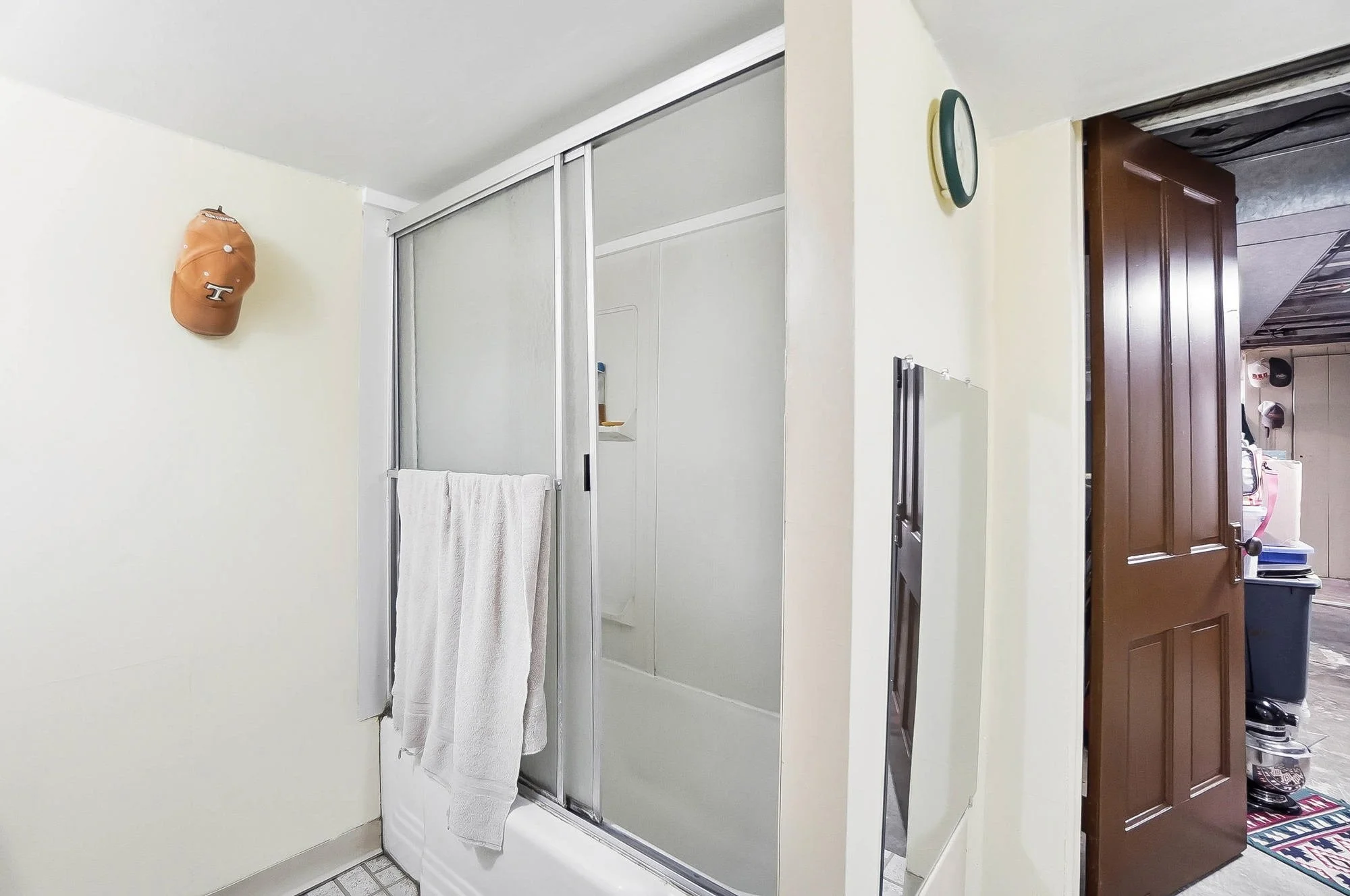 Bathroom with a glass shower enclosure, a towel hanging on the shower door, a wall clock, and a brown kitchen door partially open leading to another room.