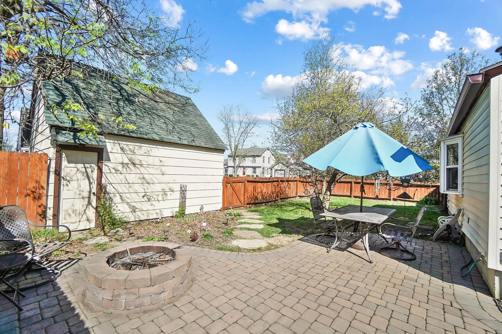 A backyard patio with a fire pit, outdoor table with chairs under a blue umbrella, a brick patio, a small garden bed, and a wooden fence, with a blue sky and some trees in the background.